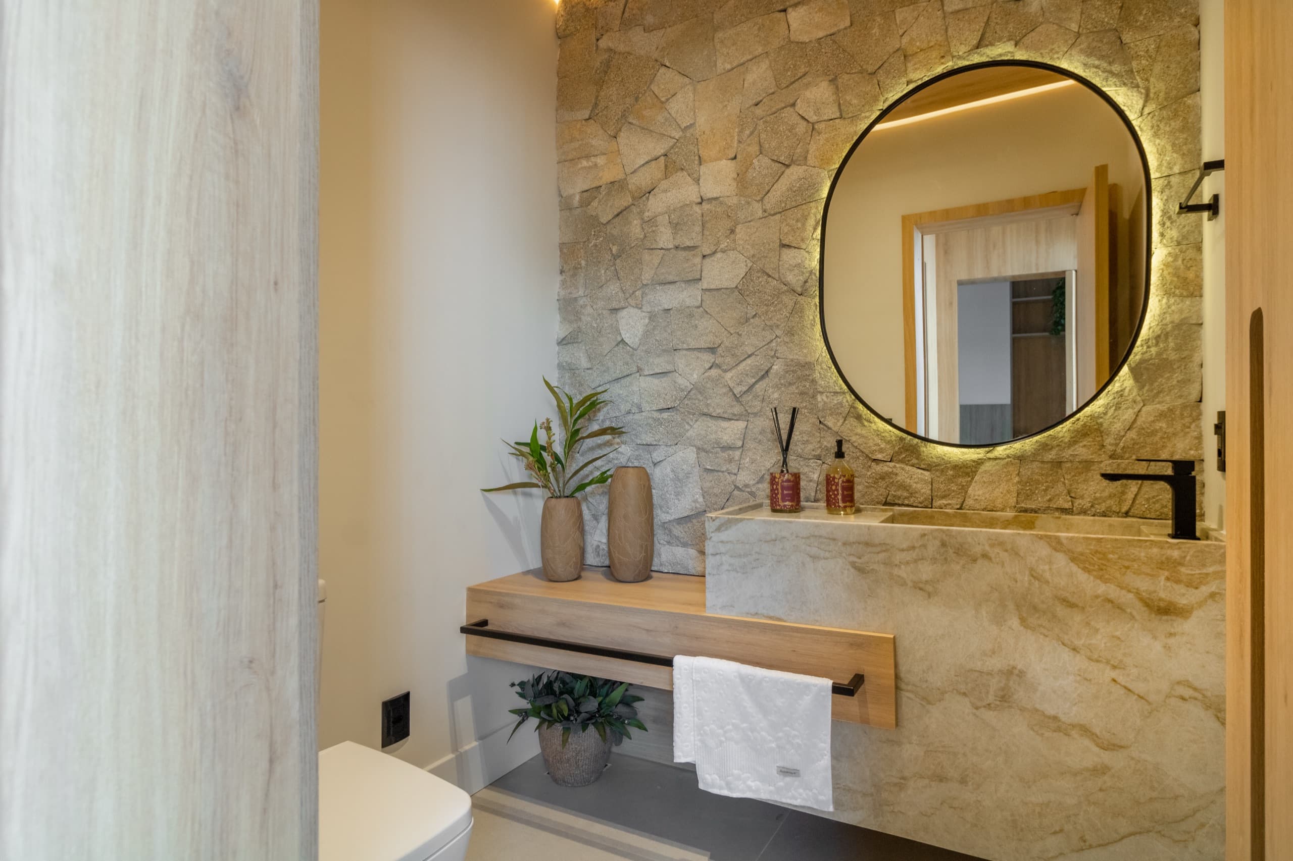 Lavabo com bancada em pedra, parede de pastilhas em relevo e espelho redondo iluminado